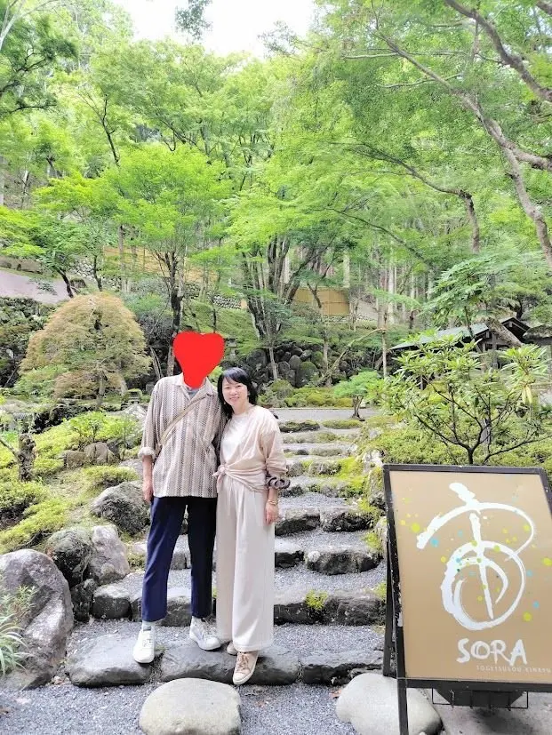 庭園の石段に立つ男女二人の写真。男性はグレーのシャツと紺色のズボン、女性はベージュのワンピースで、背景には緑豊かな木々や岩が広がる。右下には「SORA」のロゴ入り看板がある。
