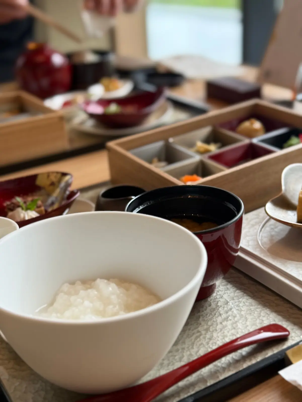 白い椀に米飯が盛りられ、赤い小鉢や木製のトレイにさまざまな料理が並ぶ和食の盛り付け