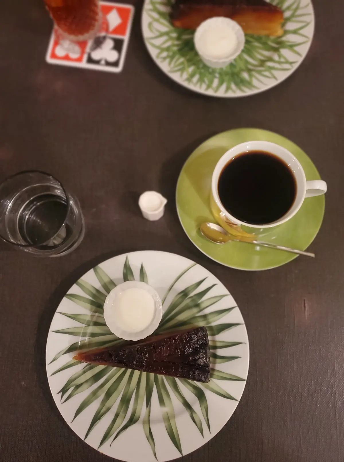 緑色の葉柄の皿に、ココア色のケーキとクリーム、コーヒーとソーサー、グラスと砂糖が置かれている様子。テーブルは暗めの茶色。