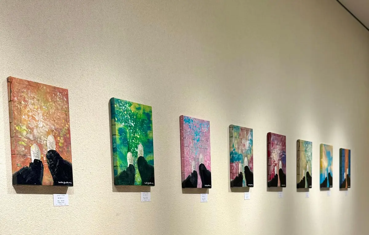 壁に沿って並んだ、抽象的な絵画の連作。それぞれに山と光の表現があり、背景はカラフルな水彩画風。絵の下には小さな白いラベルが貼られている。