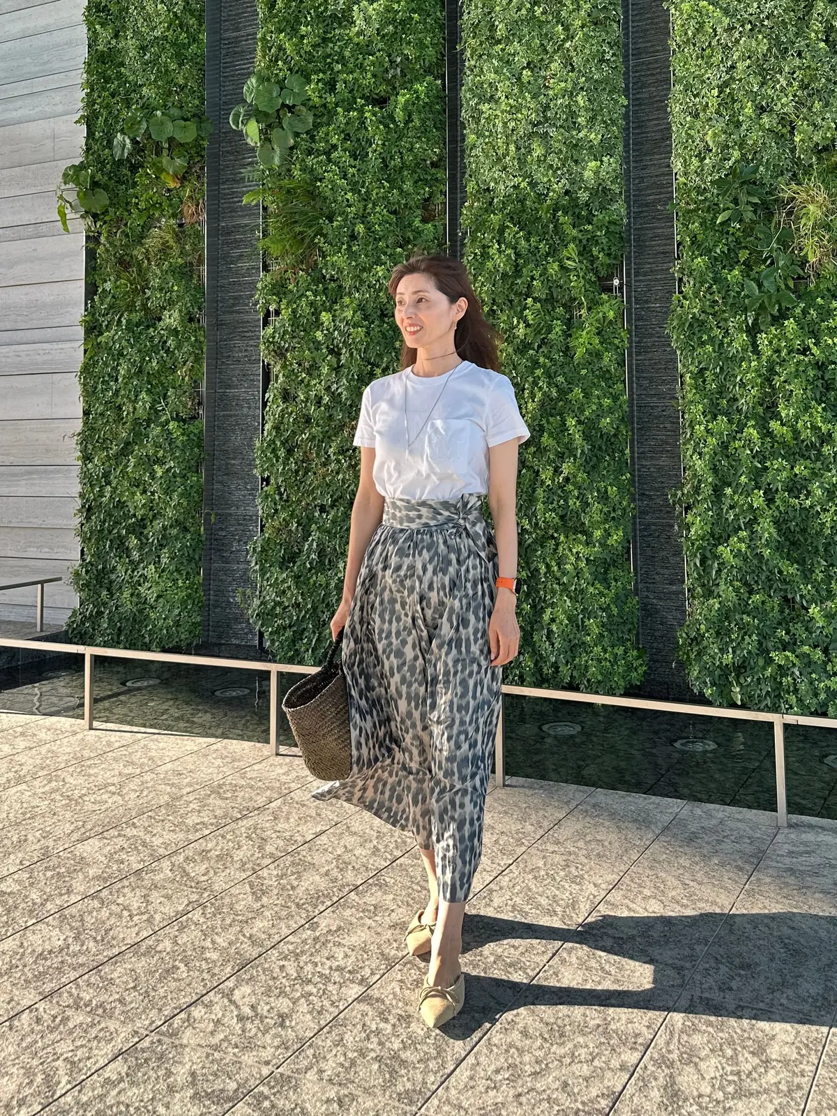 女性が白いTシャツとヒョウ柄スカートを着て、屋外の緑の壁の前を歩く。手には編みかごを持ち、笑顔でカメラに向かっている。