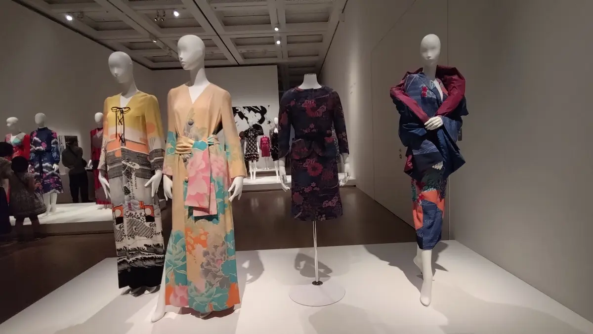 白いマネキンが並ぶ展示スペースで、様々な色や柄のドレスが展示されている。左から黄色と黒の模様のあるロングドレス、花柄のベージュのワンピース、黒のフラワープリントのミディアムドレス、青と赤のチェック柄のセットアップなどがある。