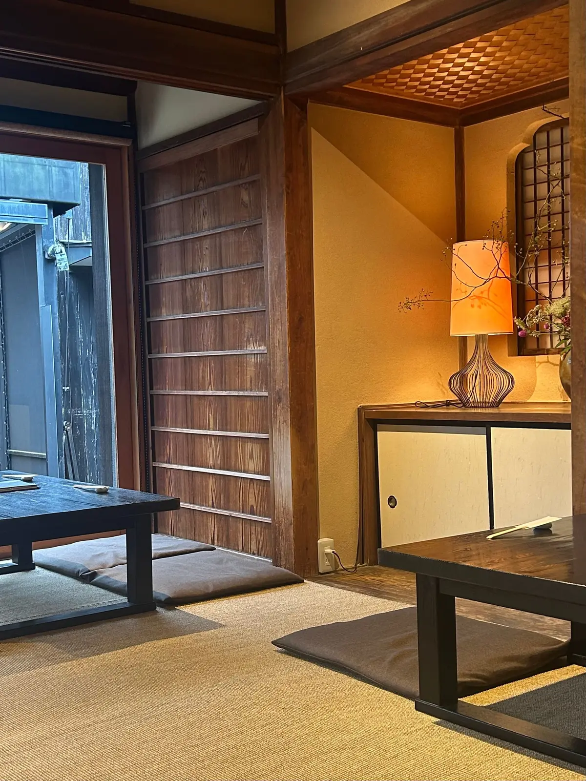 木製の障子と畳敷きの部屋。右には照明器具と装飾棚があり、左には低いテーブルが置かれている。全体的に落ち着いた茶色と黄色の色合い。