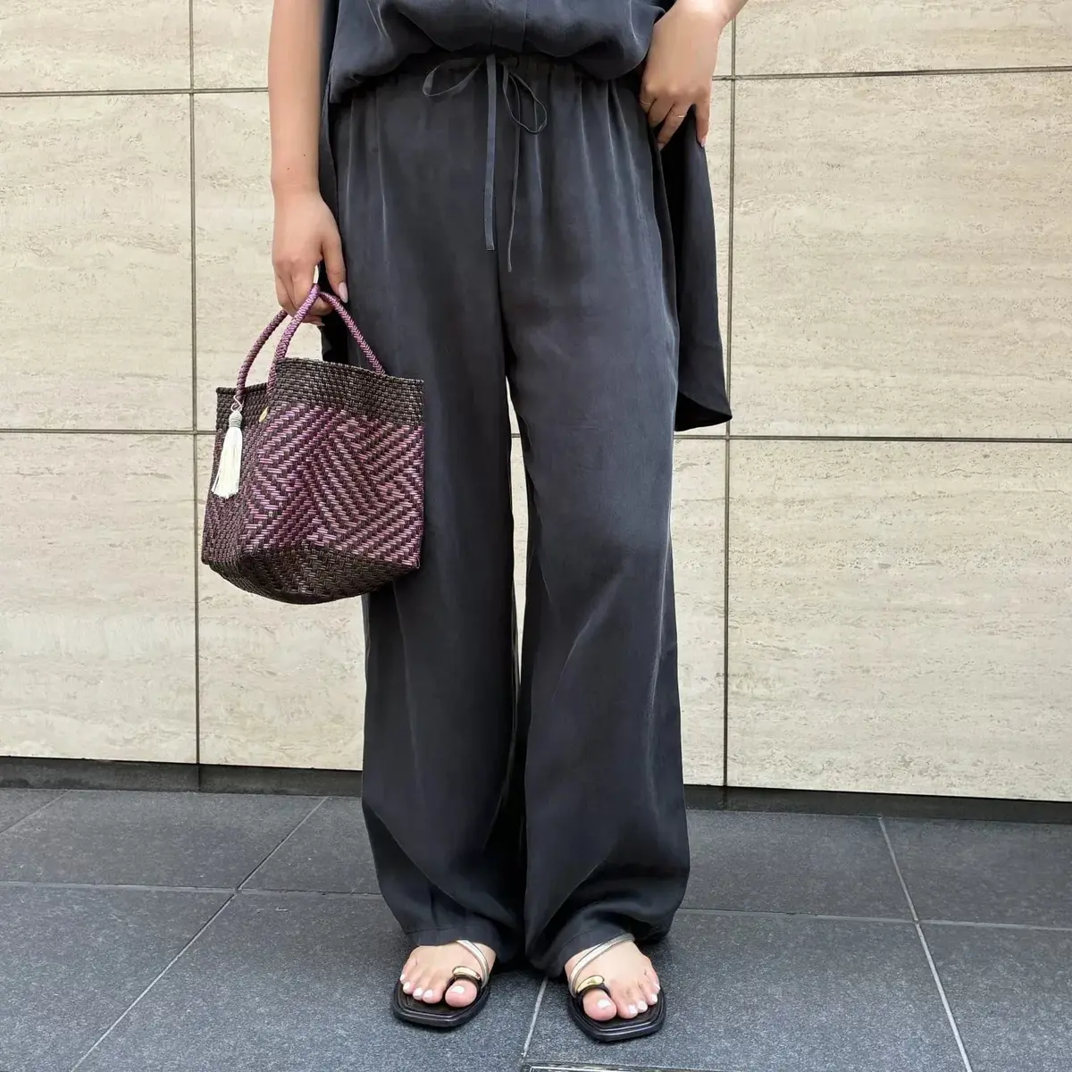 黒いワイドパンツとゴールドのサンダルを履いた女性。手には紫と茶色の編み込みバッグを持つ。背景はベージュの石壁。