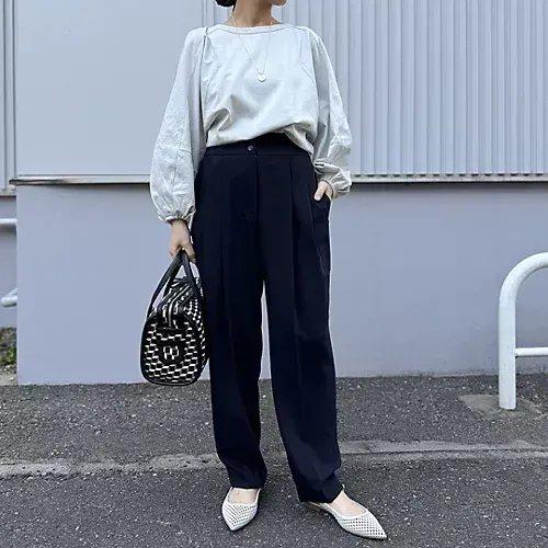 白系のトップスに濃紺のプリーツパンツを合わせた女性の全身写真。白いフラットシューズとバッグ、ゴールドのネックレスを着用。背景はグレーの建物。