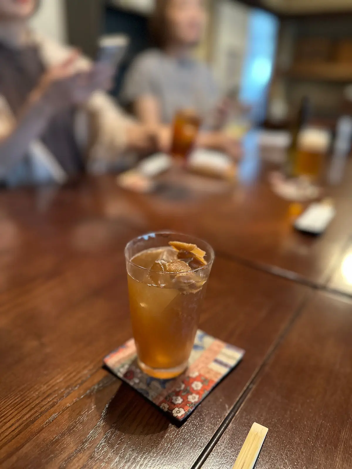 木製のテーブルに置かれた、琥珀色の飲み物。グラスにはドライフルーツのようなものが浮かび、カラフルなコースターが敷かれている。背景には人々が食事を楽しんでいる様子が見える。