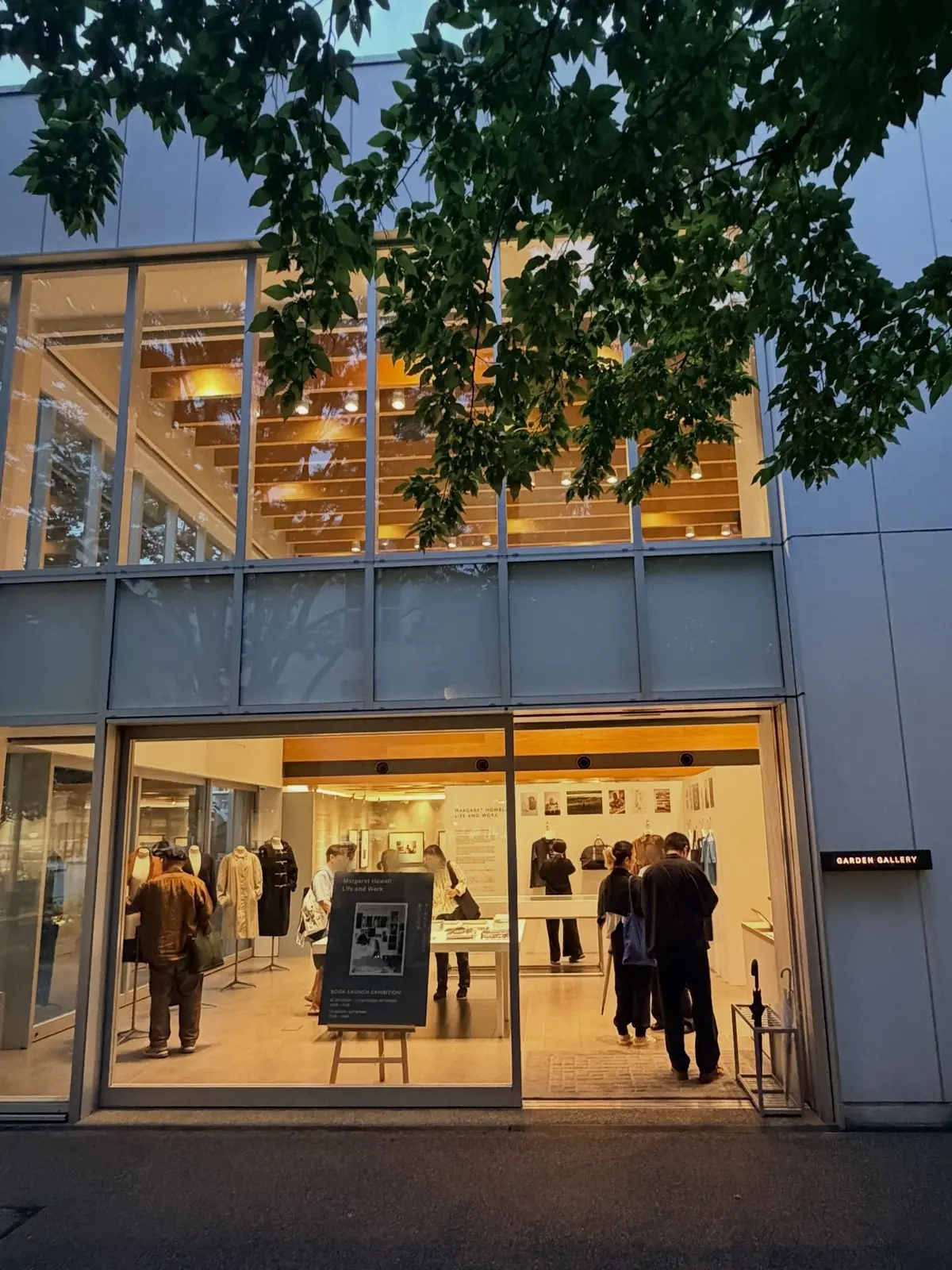 白い建物に面したガラス張りの入り口を持つ代官山「ガーデンギャラリー」。内部には展示物と見学者がおり、入り口前に展示板が置かれている。緑豊かな木が建物の上を覆っている。