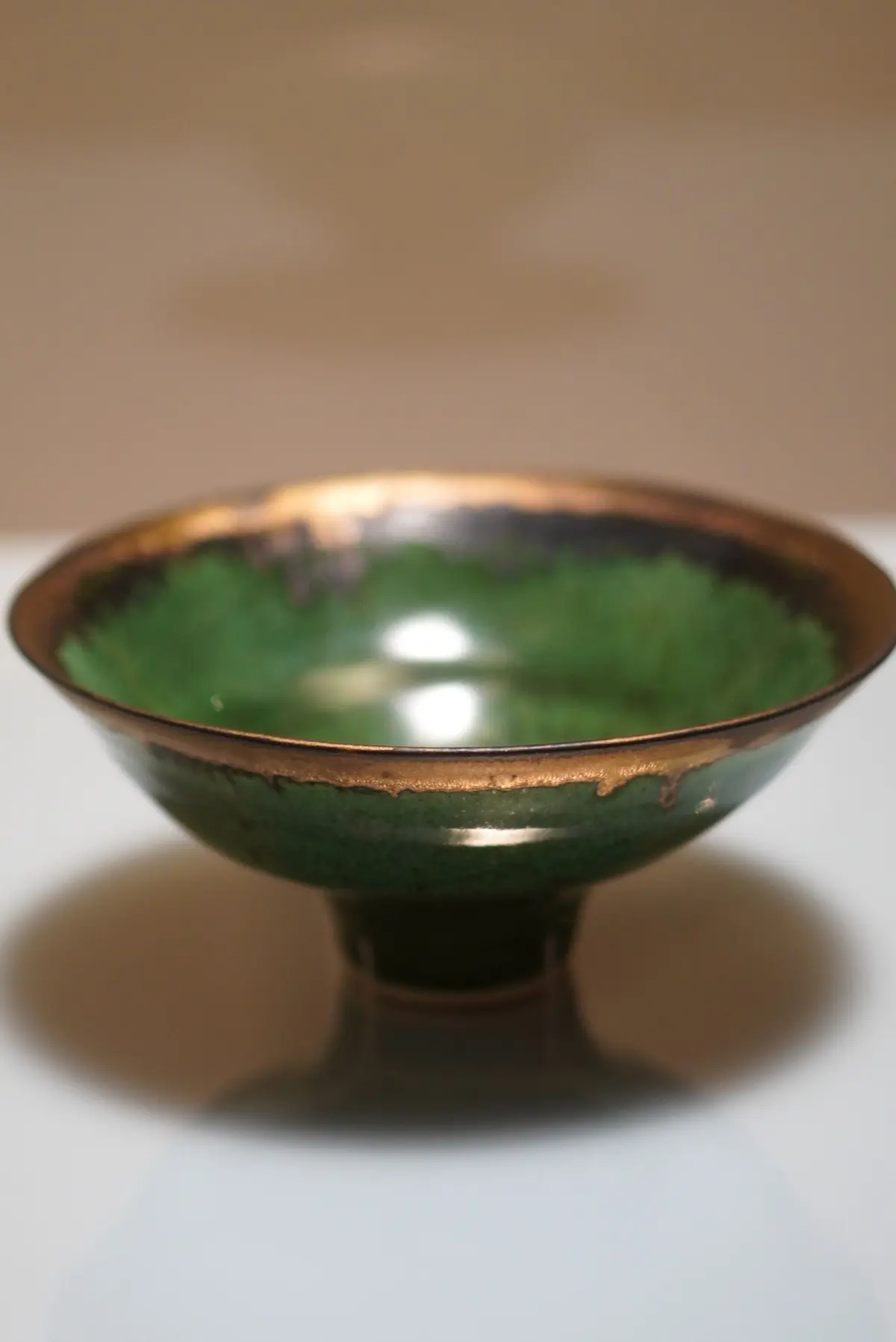 緑と金色の釉薬が施された陶器製の浅いボウル。中央に光が反射し、内側の表面が鏡のように見える。台座に置かれ、影が伸びている。