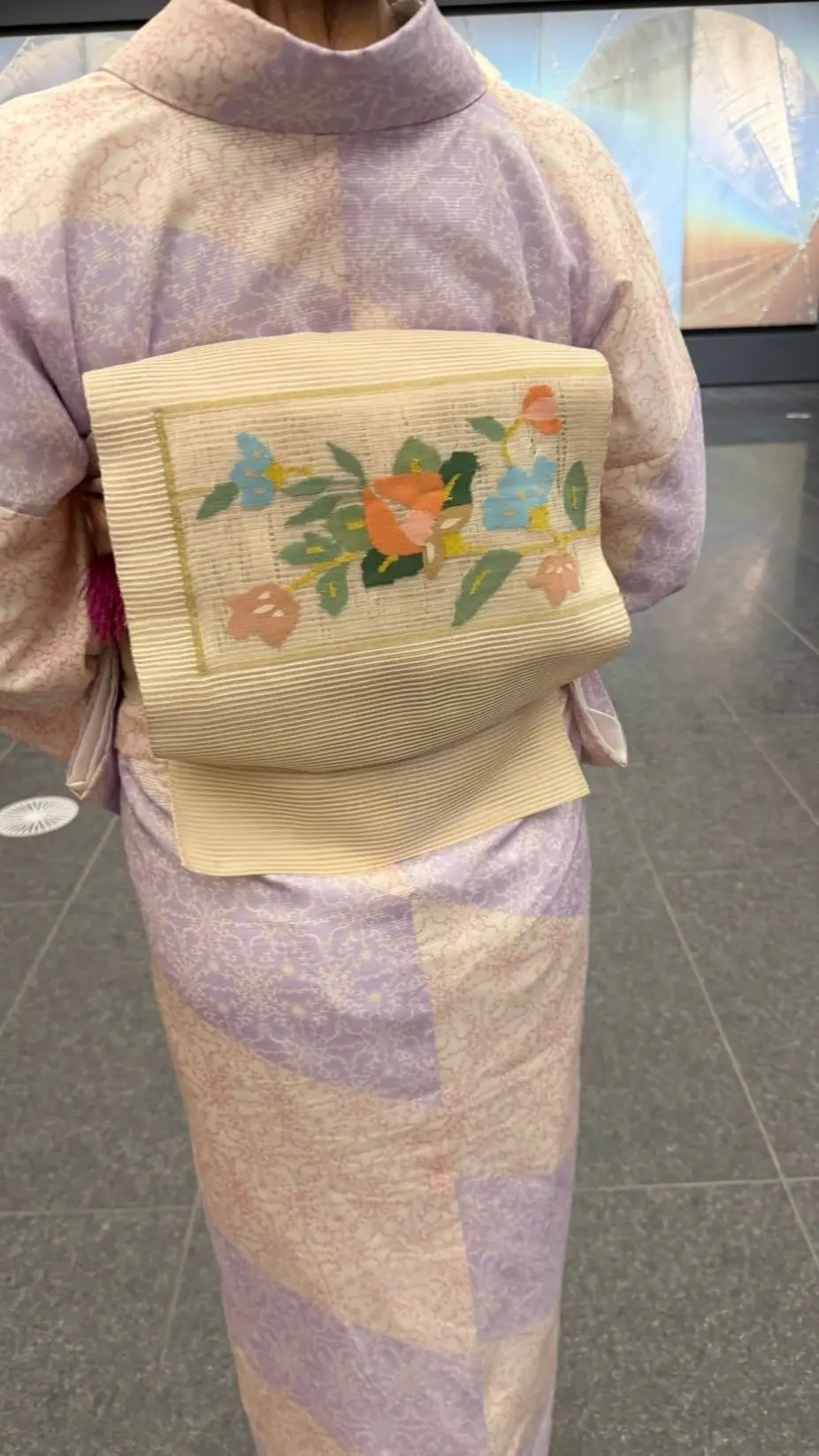 淡いピンク、白、紫のグラデーションが施された着物姿の人物。腰には生成色の帯を締め、その上に花柄の角帯が巻かれている。