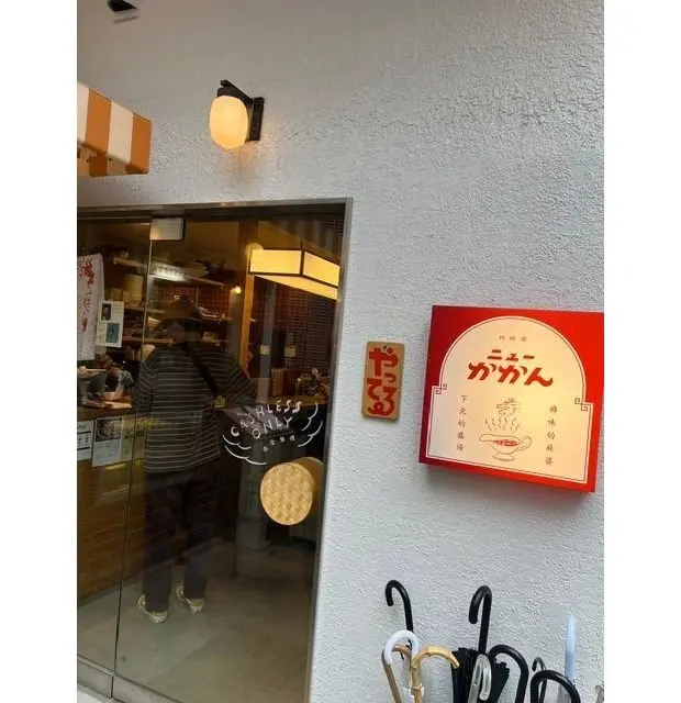 白壁の建物に設置されたガラス張りの入り口。右には赤い看板があり、日本語で店名「やなぎみ」と説明文が書かれている。入り口の窓には「キャッシュレス専用」と書かれた手書き風の文字。左には傘立てがあり、傘が並んでいる。