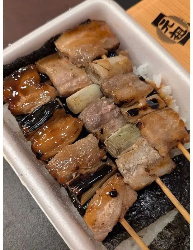 串に刺さった豚肉と野菜、ご飯が海苔で包まれたお弁当。照り焼きソースがかかっている。