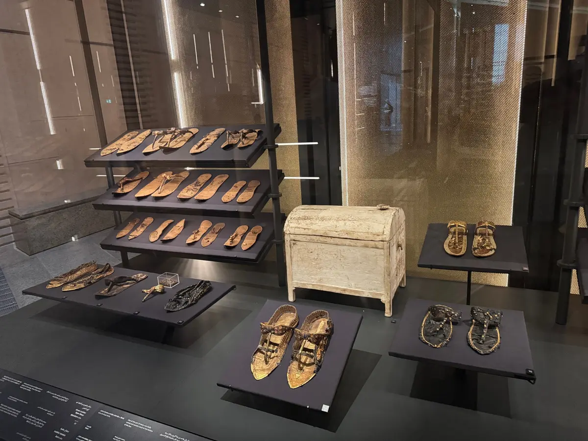 古代エジプトの展示ケース内に、金色の装飾品や靴、木製の箱が並べられている。棚や台に展示され、背景には窓からの光が差し込んでいる。