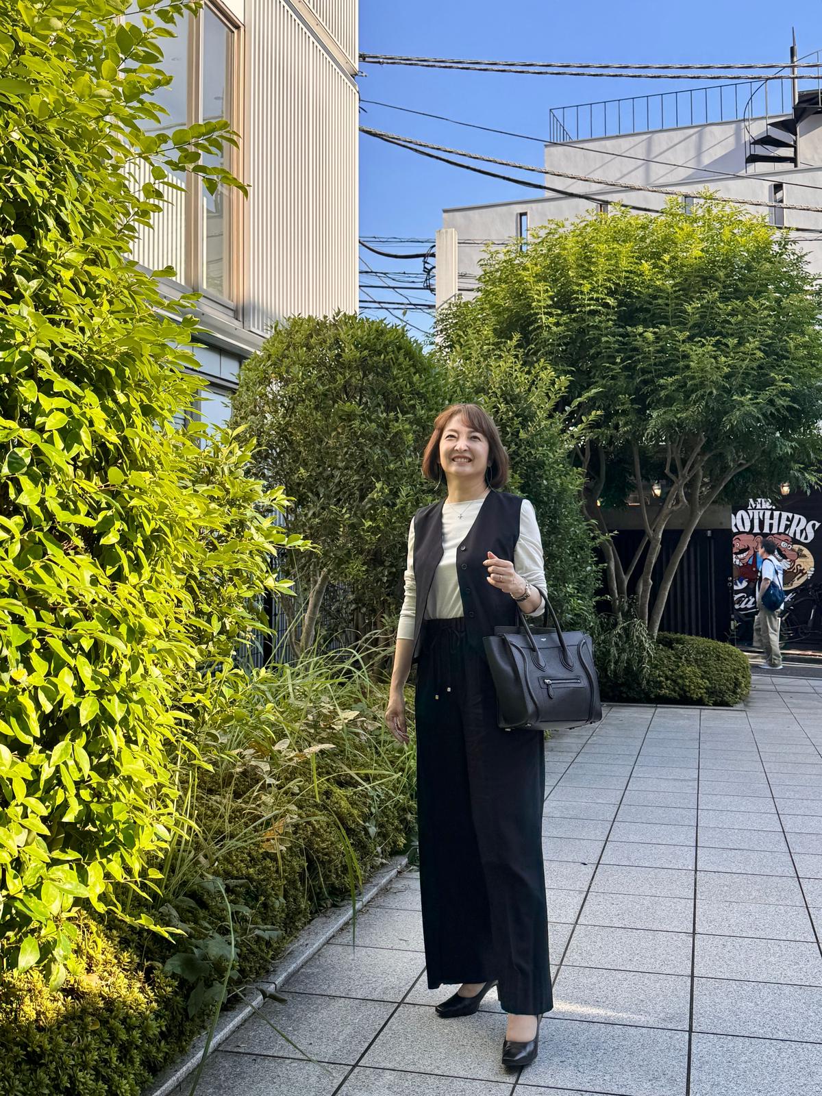 黒いパンツとベスト、白いシャツを着用した女性が、緑の植栽を背景に立っている。右手に黒いハンドバッグを持ち、グレーの靴を履いている。背景には建物と「THE BROTHERS」と書かれた看板が見える。