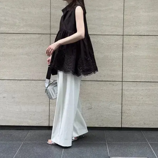 黒いギャザーのトップスと白いワイドパンツを着用した女性。右手にシルバーのバッグを持ち、横を向いている。背景はベージュのタイル壁。