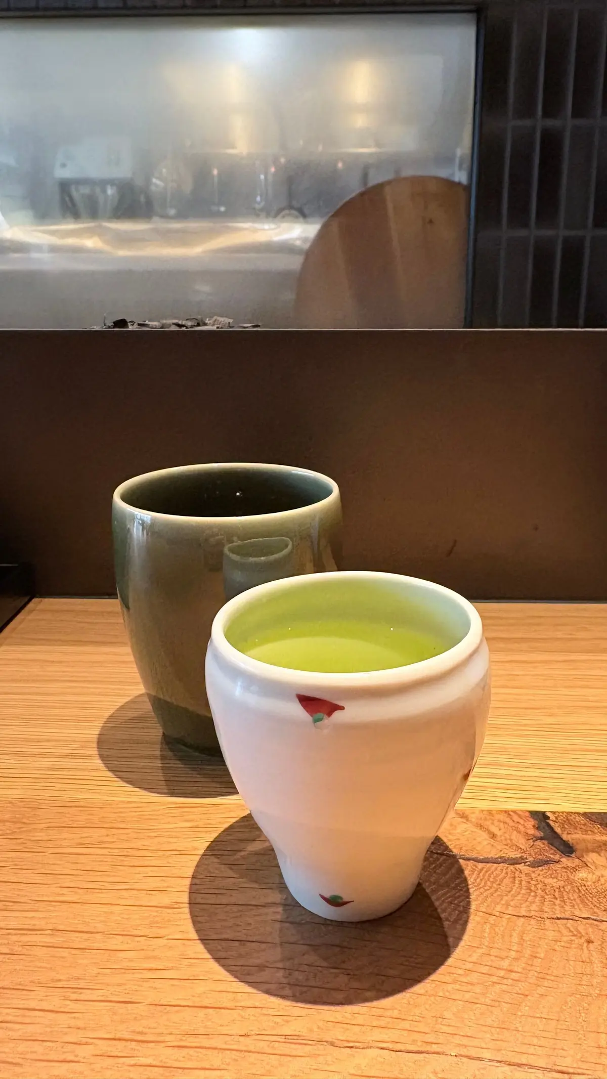 木製のテーブルに置かれた、白と緑色の茶碗2つ。片方は緑色の飲み物が入っており、もう片方は空。背景にはぼやけた厨房の様子が見える。