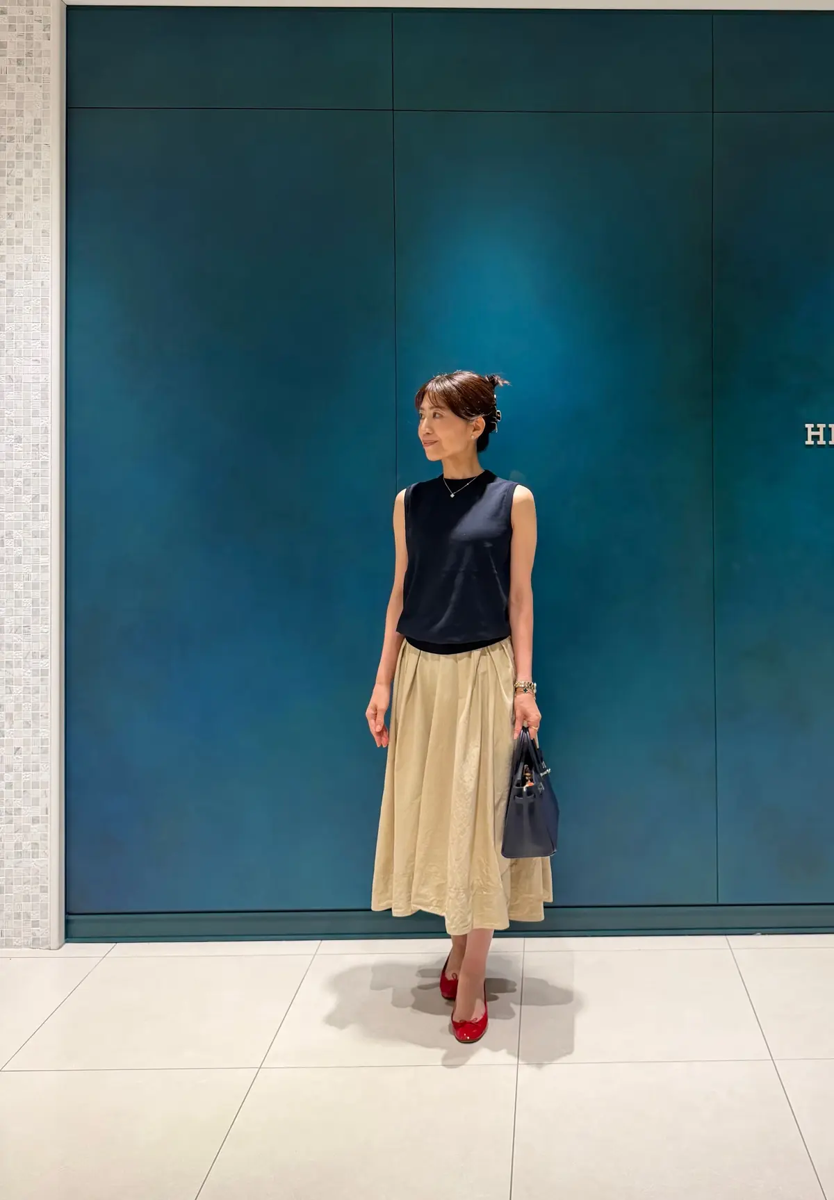 ダークブルーの壁前に、ノースリーブの黒トップとベージュのロングスカートを着た女性が立っている。赤いパンプスを履き、左手には黒のハンドバッグを持っている。