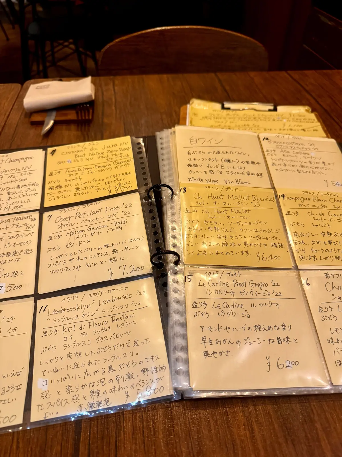 手書きの文字が書かれた2ページのメニュー。左ページはシャンパーニュやワイン、右ページは白ワインと日本酒が記載されている。各ページには商品名、説明、価格が手書きで丁寧に記載されている。背景は茶色で、手書き文字が黒色で書かれている。