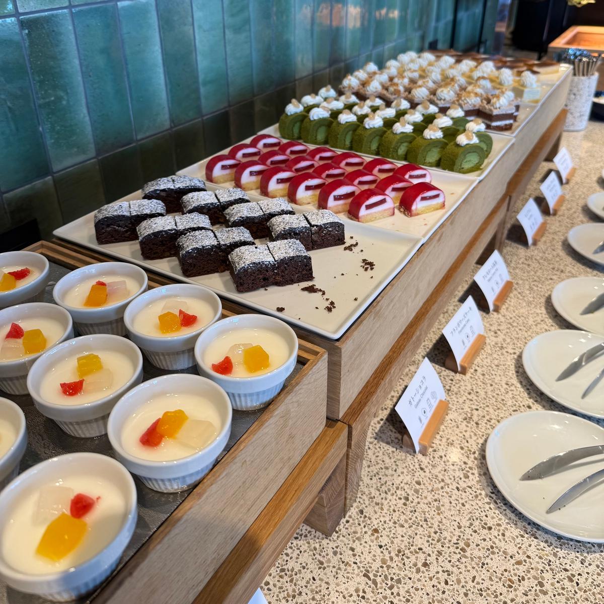 ビュッフェ台に並んだ様々なデザート。ロールケーキ、抹茶ケーキ、ブラウニー、カップに入ったプリンなどが並び、白い皿とカトラリーが置かれている。