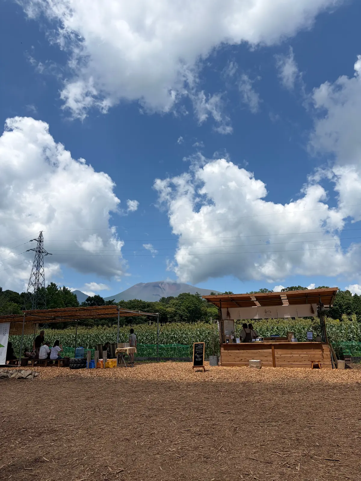 青空に白い雲が広がり、遠くには山が見える風景。手前には土の地面に木製の小屋と、緑色のトウモロコシ畑が広がる。