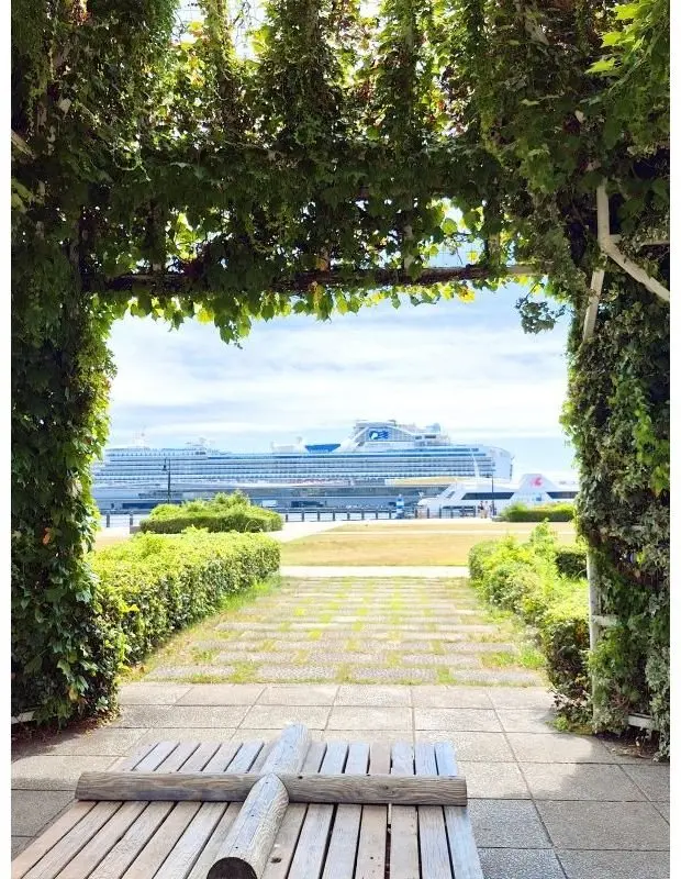 緑のツタで覆われたアーチの向こうに、大型の白い客船が停泊している風景。手前には木製のベンチと、芝生と生垣が配置された歩道が続く。背景には雲が浮かぶ青空。