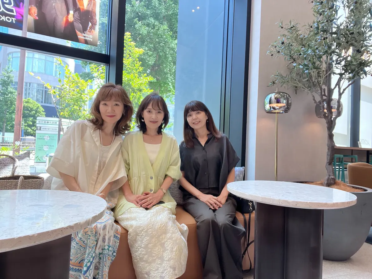 カフェのような空間で、3人の女性が並んで座っている写真。左から、ベージュのシャツを着た女性、淡い緑色のワンピースを着た女性、黒いシャツとズボンを着た女性。それぞれ笑顔でこちらを見ている。背景には窓から外の景色が見える。