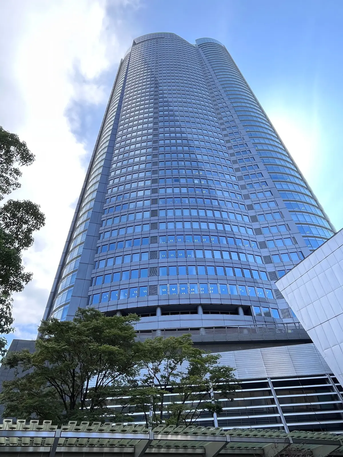 高層ビルが青空高くそびえ立つ。窓が規則的に並び、建物は曲線的な形状。手前には緑の木々が生い茂り、白い雲が浮かぶ。