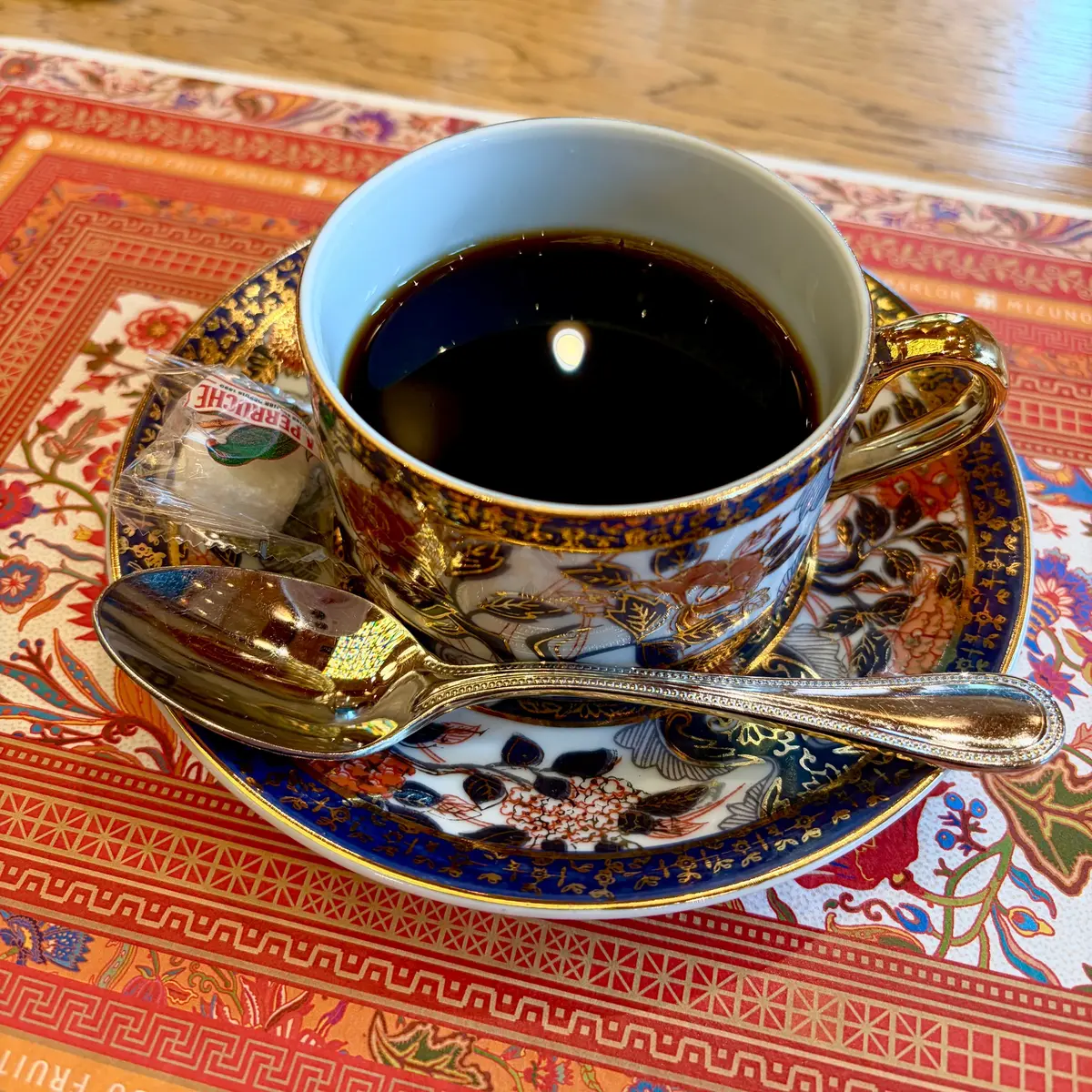 青いマグカップに黒いコーヒーが入っており、金の装飾が施されたプレートに置かれています。プレートには花と鳥の模様があり、銀のスプーンが横に置かれています。背景には赤と金の模様のあるテーブルクロスが広がっています。