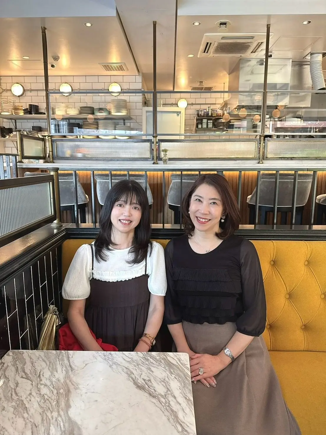 レストランのブース席で、笑顔の若い女性2人が並んで座っている。左の女性は白いブラウスに茶色のワンピース、右の女性は黒のトップスにグレーのスカートを着用。背景には白いタイルと厨房が見える。