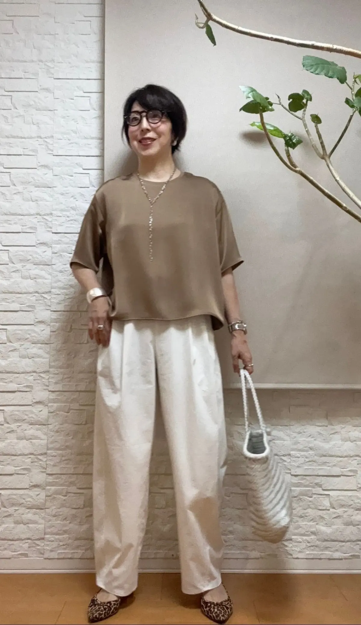 女性がベージュの半袖シャツと白いワイドパンツを着用し、壁に寄りかかっている。右手には白い編みかごバッグを持ち、左手は腰に置いている。壁は白いレンガ調で、右側には緑の葉を持つ植物が配置されている。