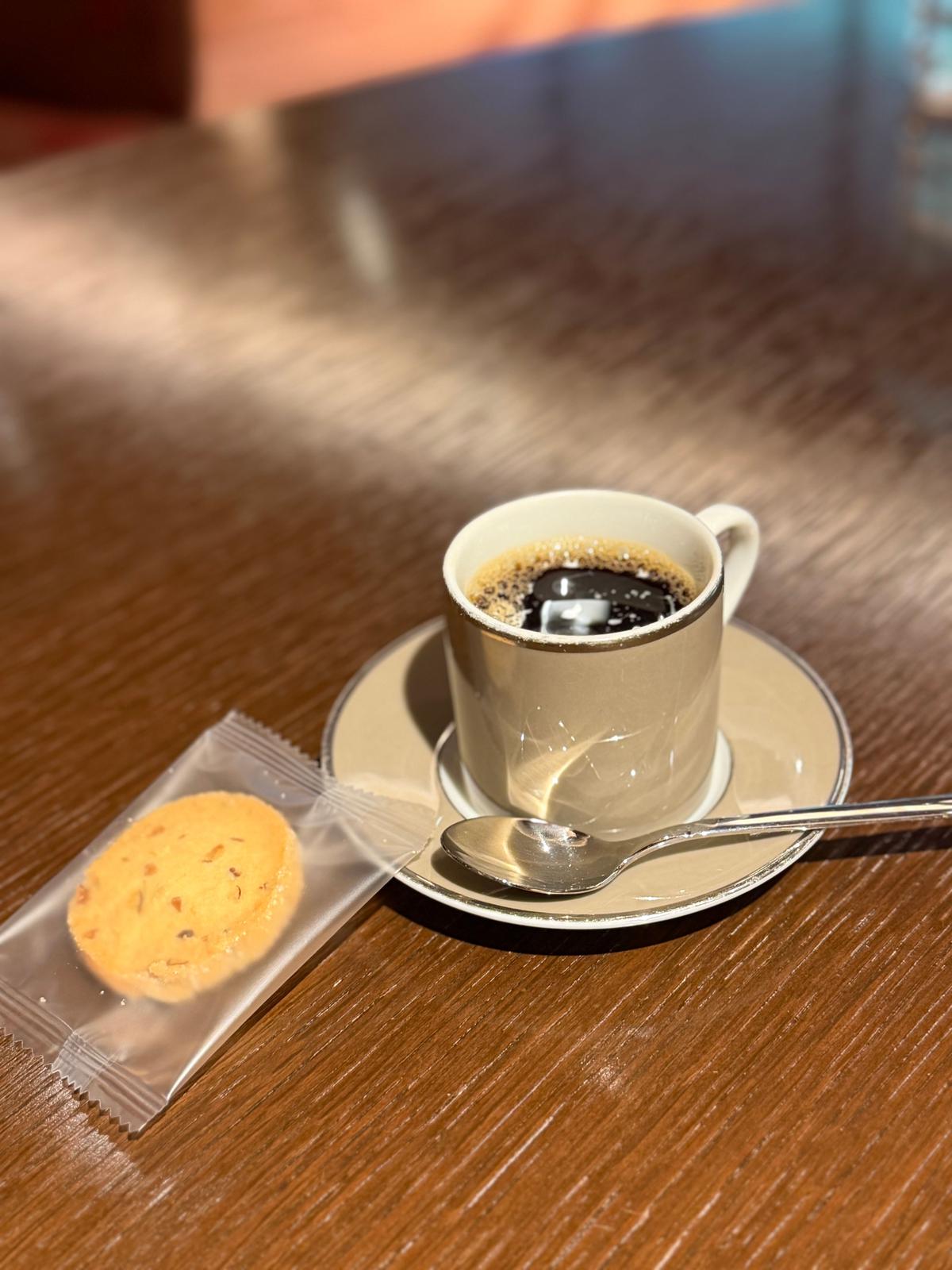 木製のテーブルに置かれたコーヒーカップとクッキー。カップはベージュ色で、ソーサーとスプーンが添えられている。クッキーは透明な袋に入っている。