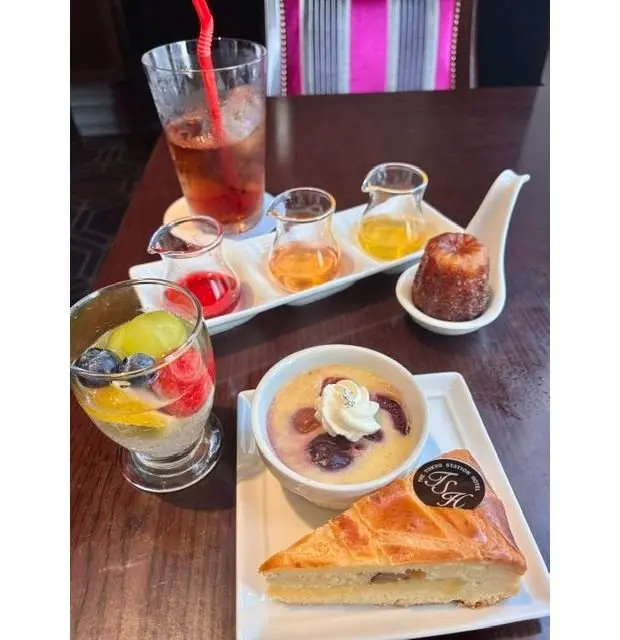 木製のテーブルに置かれた、様々なデザートと飲み物の盛り合わせ。左上にはアイスティー、左下にはフルーツゼリーとカットフルーツ、中央にはカスタードプリンとチーズケーキ、右にはマカロンとミニャルディーズ。
