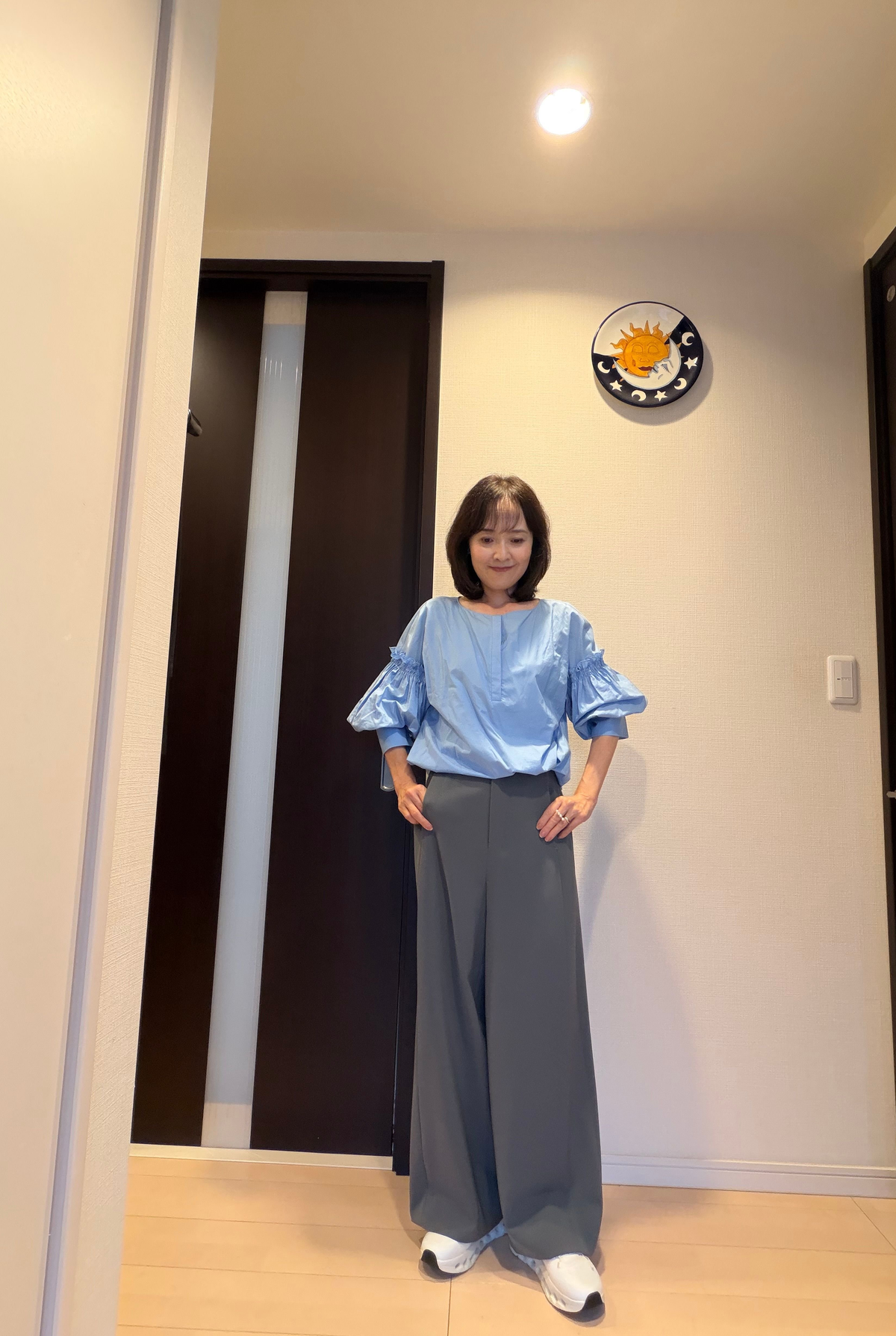 青色のパフスリーブブラウスとグレーのワイドパンツを着用した女性が立っている。
