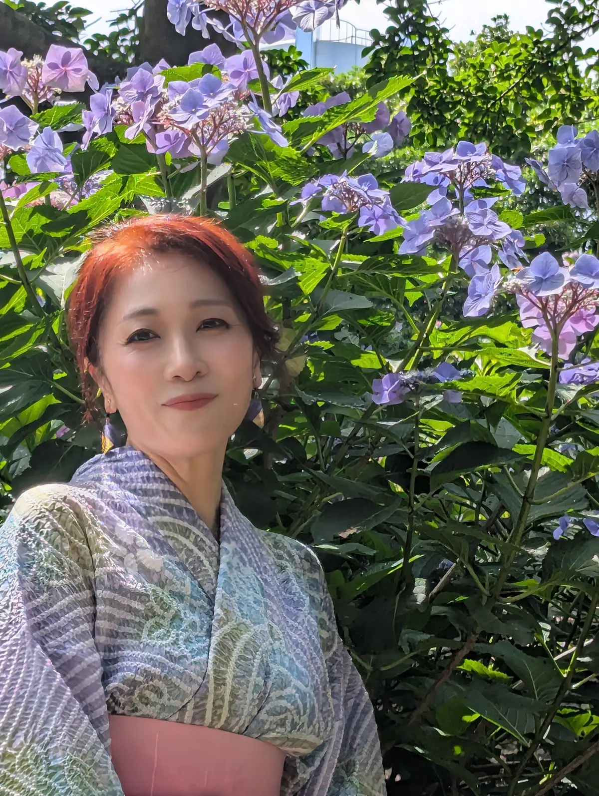 赤い髪をした女性が青紫の花々の中央に立っており、青緑の和服を着ている