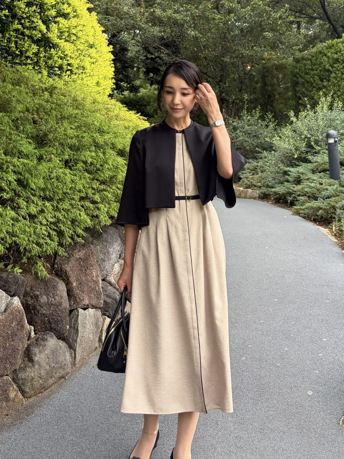 ベージュのミモレ丈ワンピースに黒のボレロを羽織った女性。右手に黒いハンドバッグを持ち、公園の遊歩道に立っている。上品な装いと落ち着いた表情が印象的。