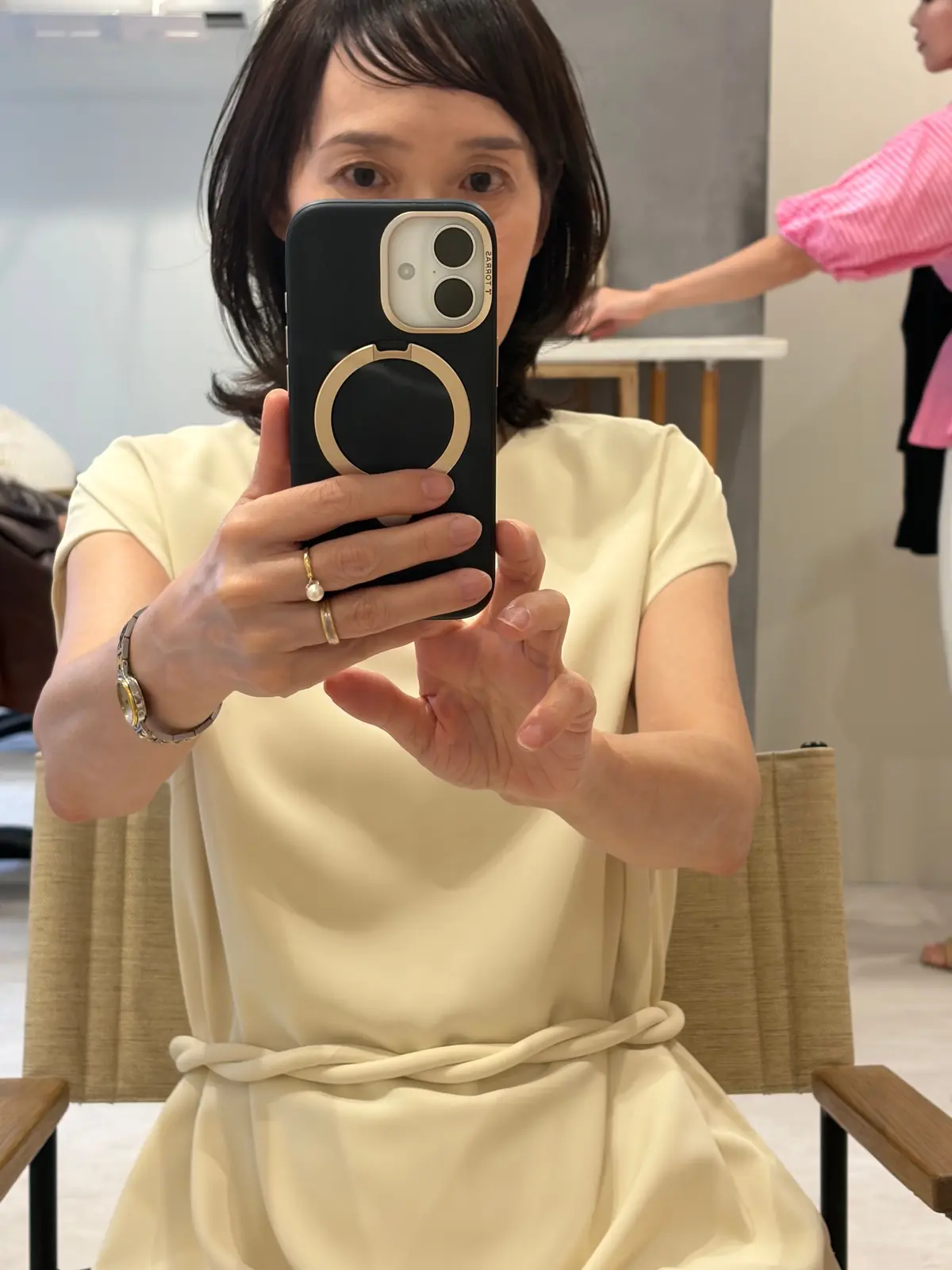 黄色のワンピースを着た女性がスマートフォンで自撮りをしている。茶色のボブヘアで、左手には指輪をはめている。