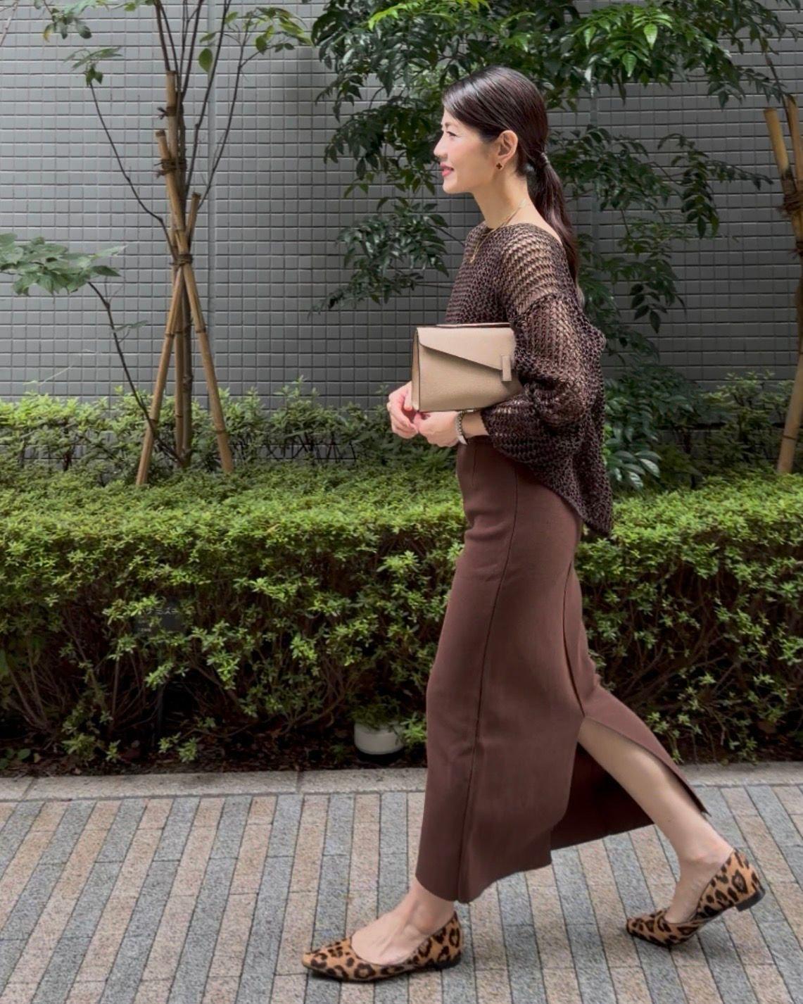 茶色のスカートと網目状のトップスを着た女性が歩いている。レオパード柄のフラットシューズを履き、ベージュのハンドバッグを持っている。背景には緑の植栽と灰色の壁がある。