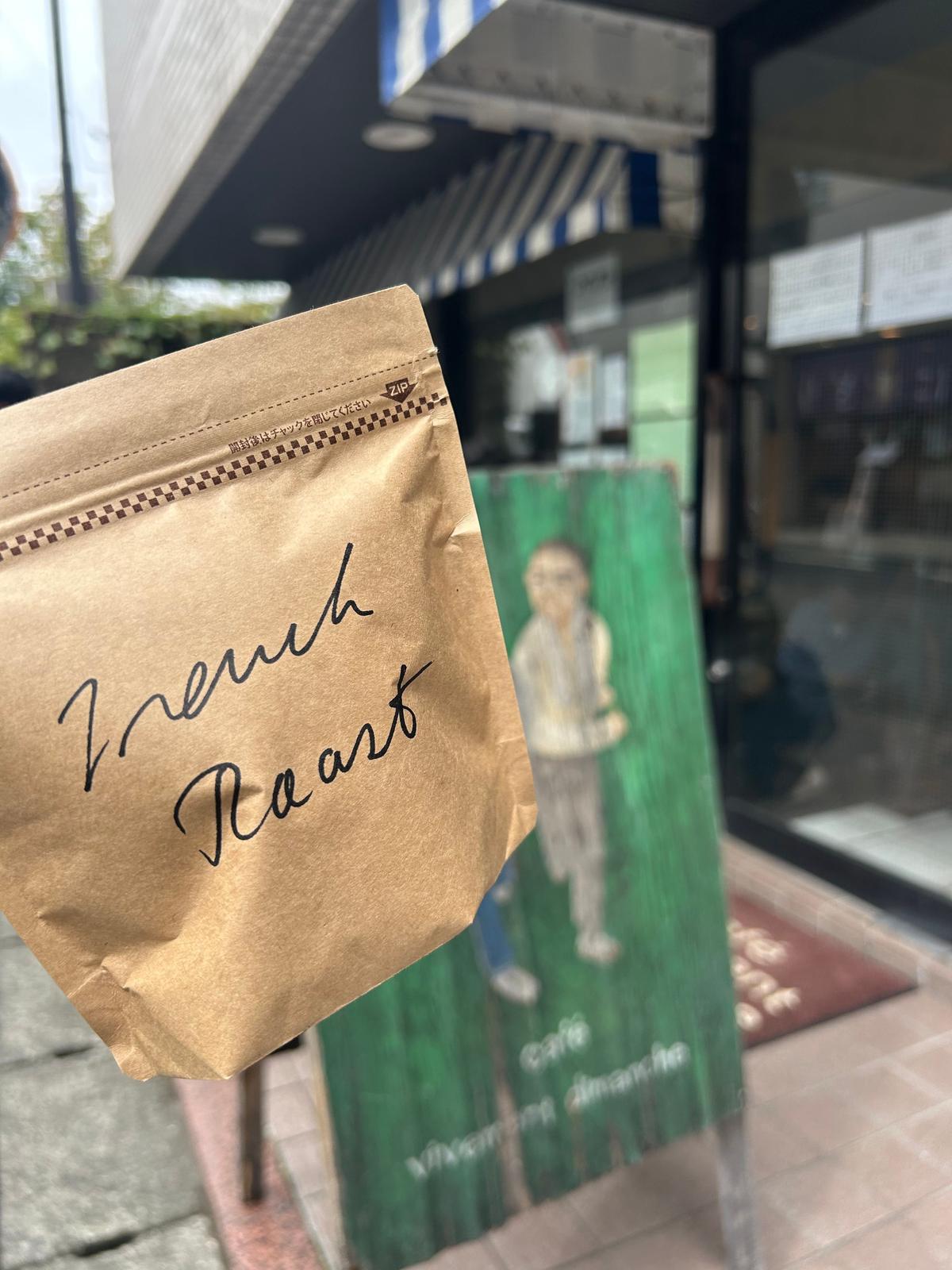 茶色の紙袋に「French Roast」と手書き風の文字で書かれたコーヒー豆の袋。背景にはカフェの窓と、人物が描かれた看板が見える。