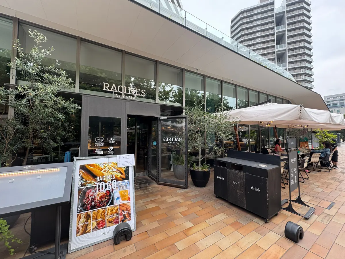 RACINES AOYAMAのテラス席、屋外のテーブルと椅子、傘付きの席、植物、メニュー看板が並ぶ
