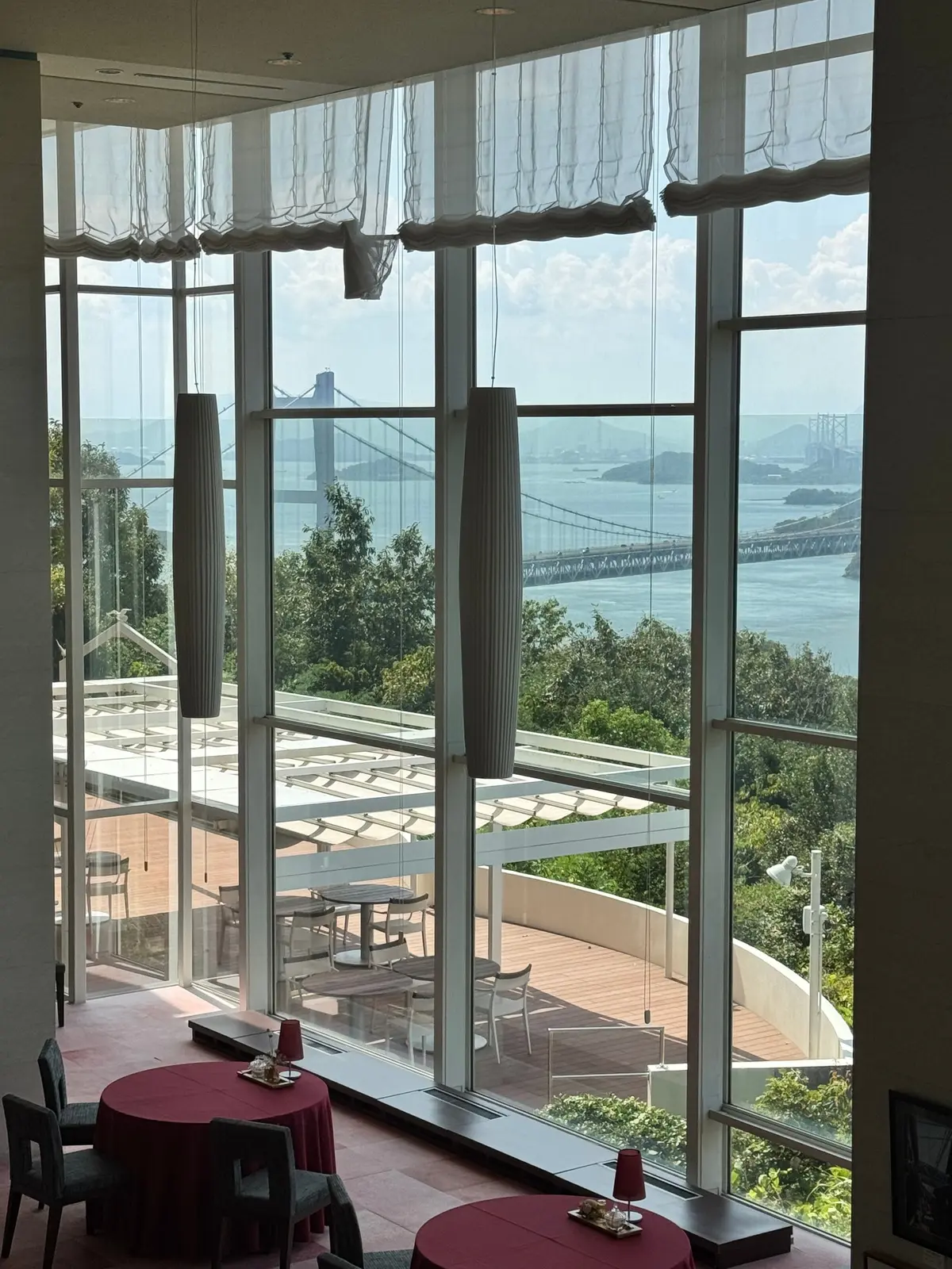 高層階のレストラン内、大きな窓から橋と水辺の景色が見える。窓には白いカーテンがかかり、窓辺には赤いテーブルと椅子が配置されている。