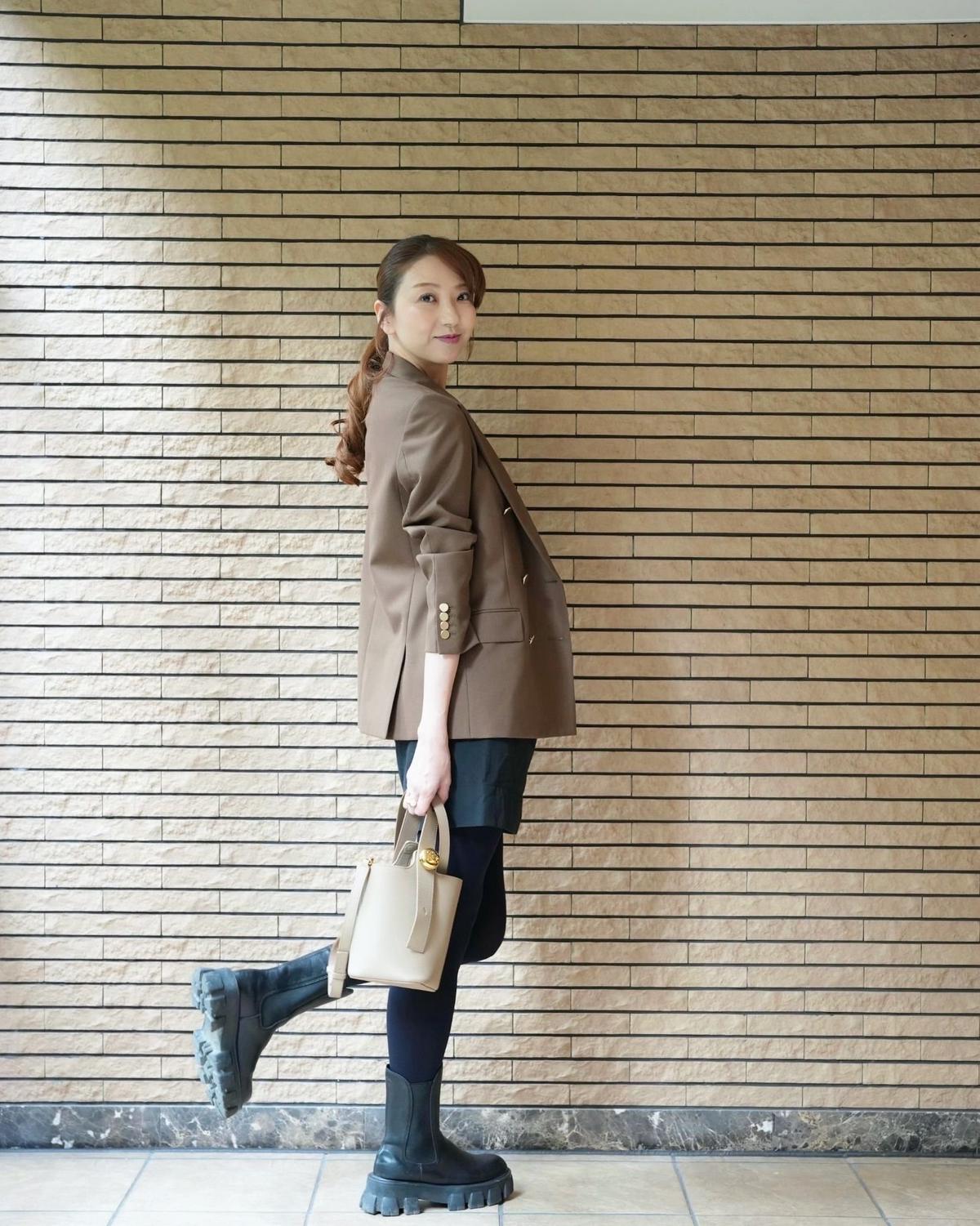 茶色のジャケットに、紺色のショートパンツとタイツ、黒い厚底ブーツを合わせた女性。右手にオフホワイトのハンドバッグを持ち、振り返ってこちらを見ている。背景はベージュのタイル壁。