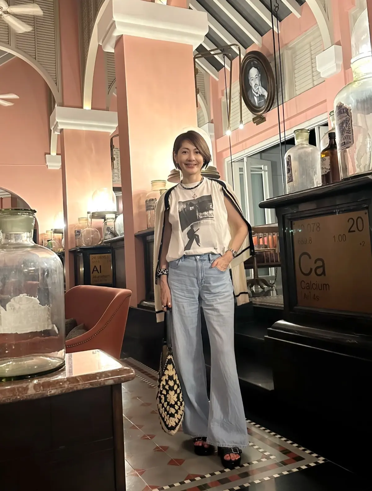 ピンクの壁とアーチ型の装飾が特徴的な空間に立つ女性。白いTシャツに裾の広げたデニムを着用し、黒いサンダルを履いている。背景にはガラス容器や周期表のポスターが見える。