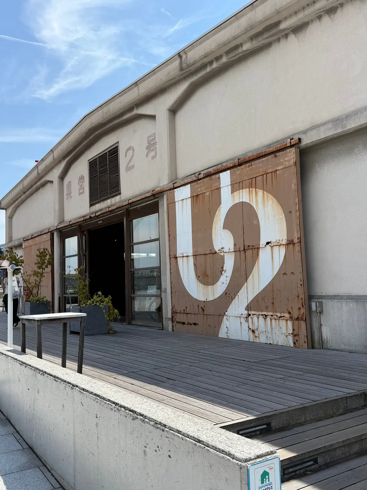 白い壁に大きな「ひ」の文字が描かれた建物の外観。右側には錆びた金属製の板が貼られ、左側には窓と「2号」の文字が見える。木製のデッキが設置され、白いテーブルと椅子が置かれている。青空が背景。尾道、ONOMICHI U2。デニムの街で尾道観光、広島。
