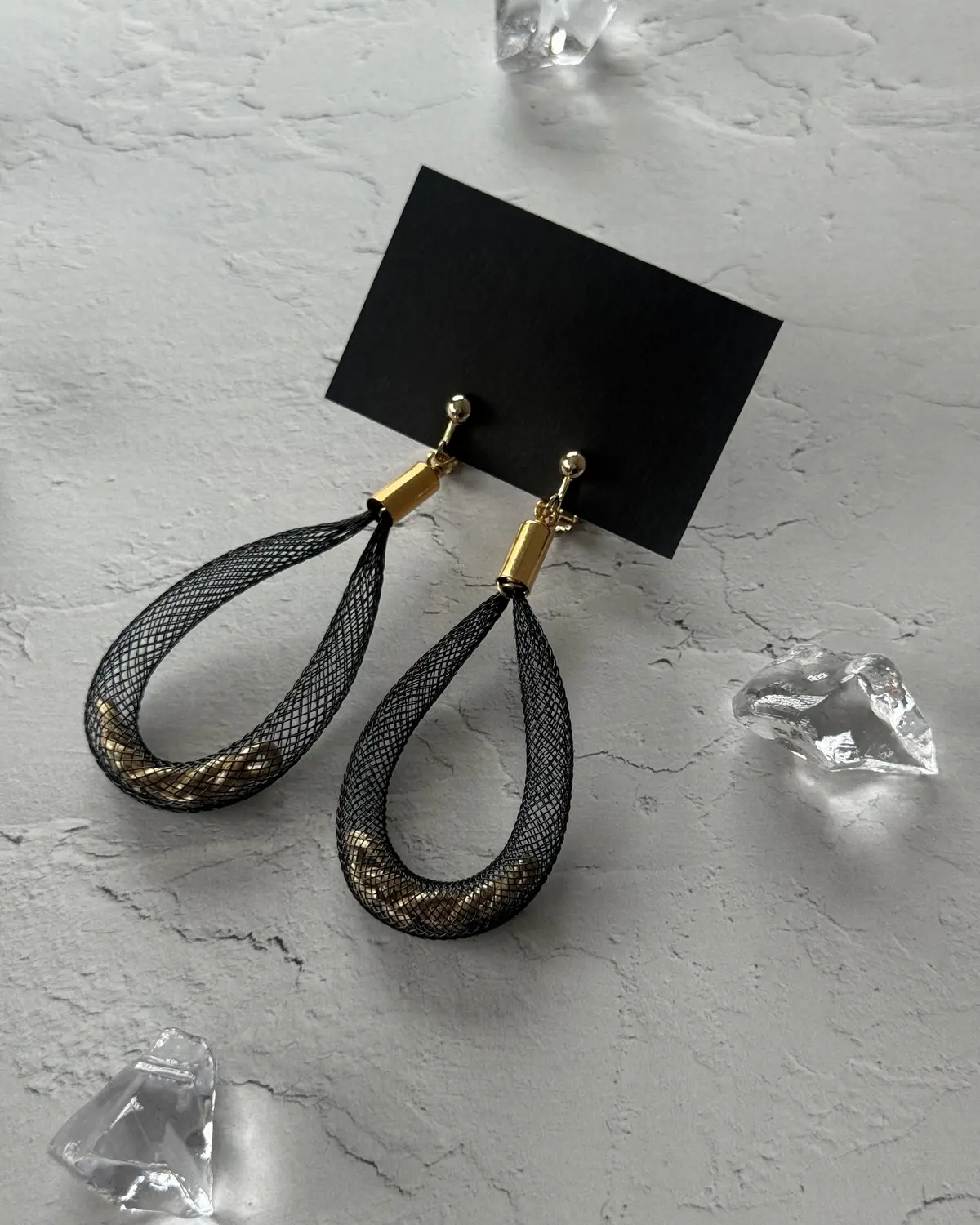 黒いメッシュ素材と金色のワイヤーでできた、左右対称のドロップ型のピアス。背景には黒いカードと透明な結晶が散らばる、灰色がかったテクスチャの表面。