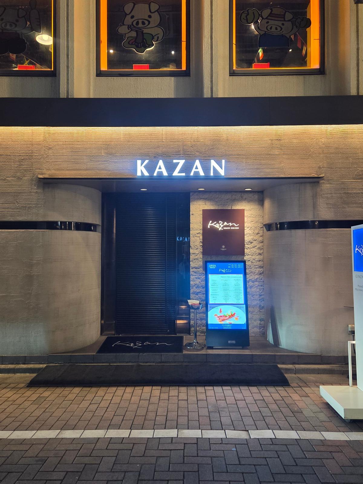 夜の街並みにある「KAZAN」と表示されたレストランの入り口。黒いドアと石積みの壁、デジタルメニューボードが設置されている。上部には、豚のイラストが描かれた窓がある。