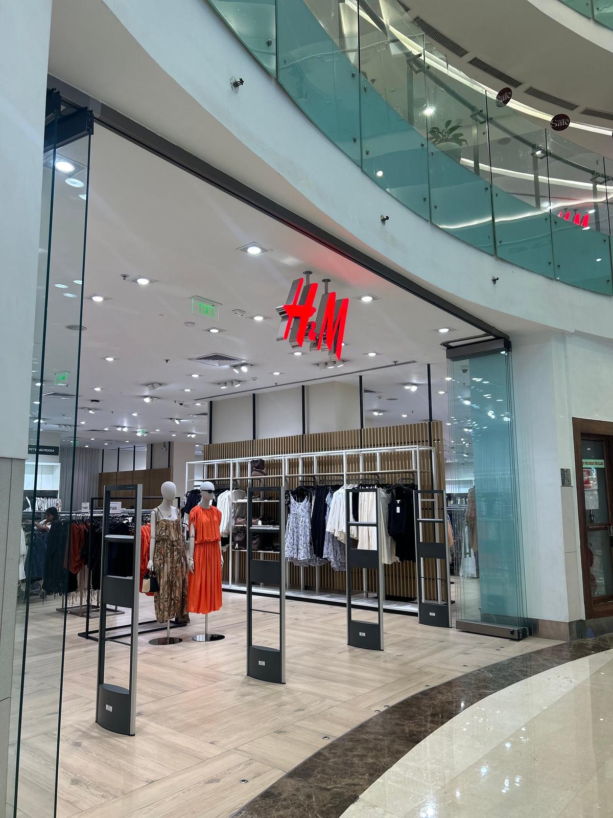 ショッピングモール内にあるH&Mの店舗。ガラス張りのショーウィンドウに、 mannequinsが様々な服を着けて展示されている。背景には、白い壁と天井、吹き抜けの構造が見える。