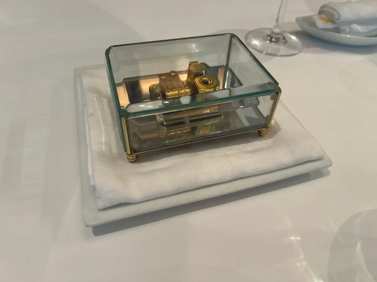 ガラスケースに入ったゼンマイ仕掛けの機械。真鍮製の部品が複雑に組み合わされており、レトロな雰囲気を醸し出している。白い布の上に置かれ、テーブルに配置されている。