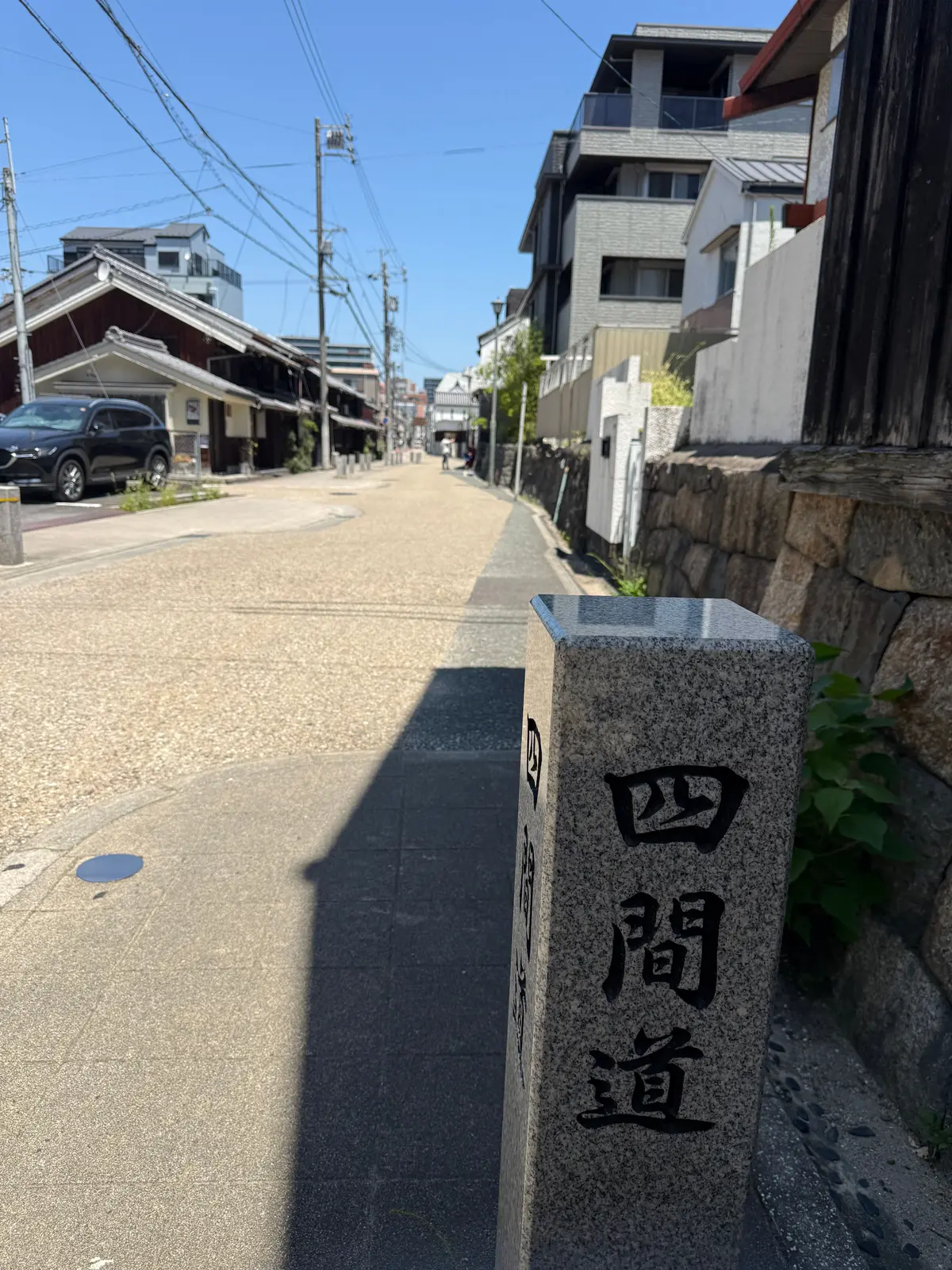 石柱に「四間道」と刻まれた、日本の住宅街の風景。細い道が遠くまで続き、左右には伝統的な家並みと現代的な建物が並ぶ。青空の下、影が長く伸びている。