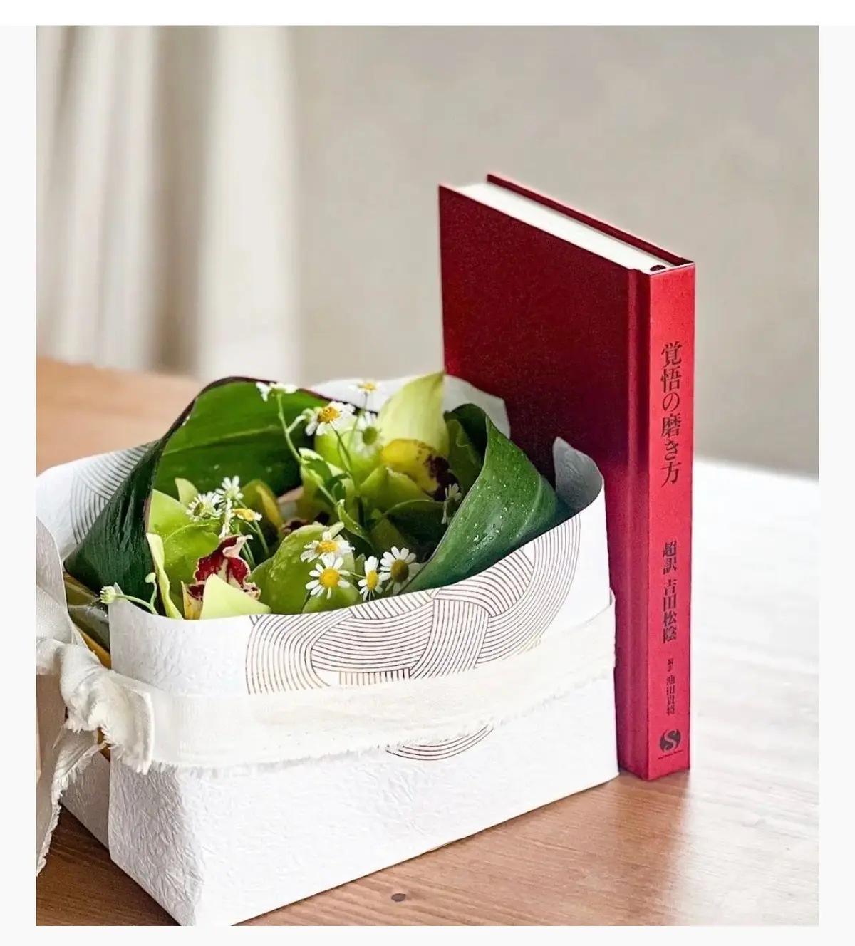 白い箱に花束が添えられ、赤い本の隣に置かれている。本は縦に立っており、タイトルが書かれている。
