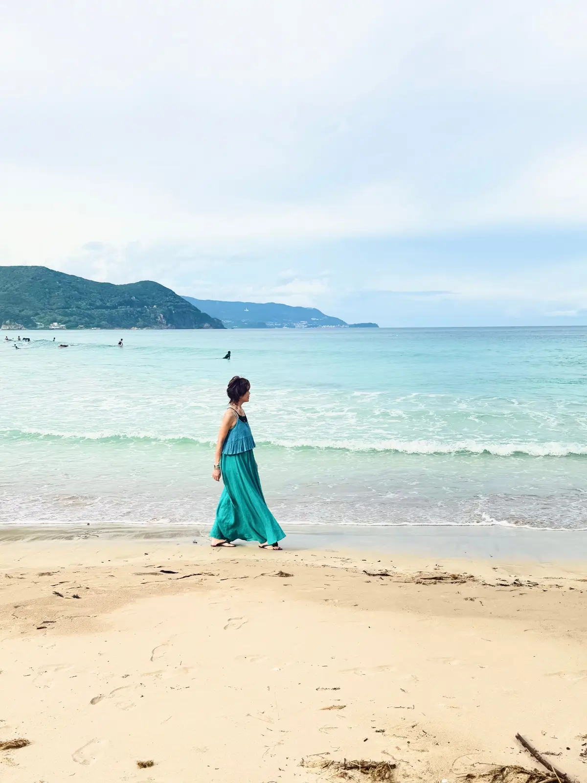 海辺を歩く女性。鮮やかなターコイズブルーのワンピースを着て、右方向に視線を向けている。背景には緑の丘と青い海、白い空が広がる。砂浜には人の足跡が残っている。