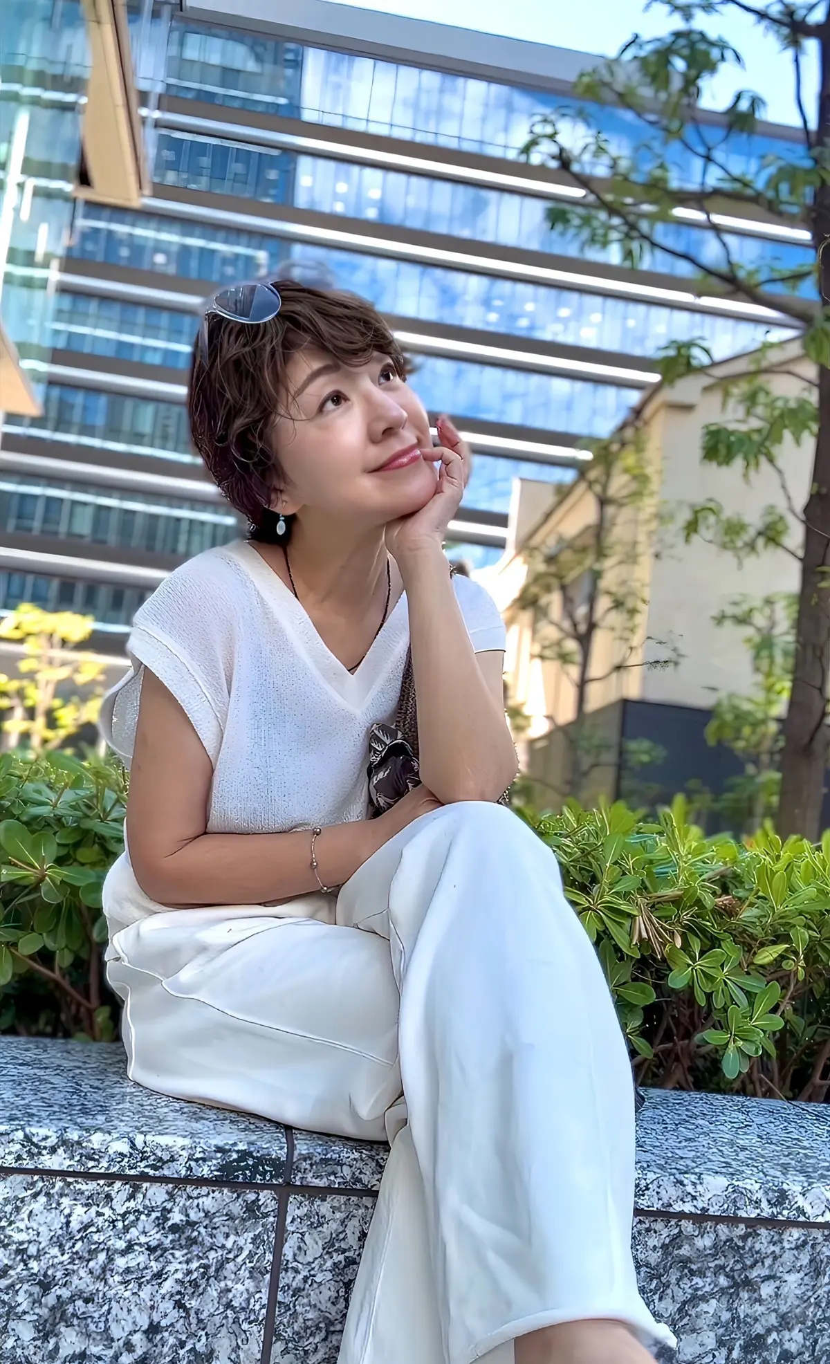 白いトップスとパンツを着た女性が、石の縁に腰掛け、遠くを見つめている。茶色の短髪で、黒いサングラスを頭にかけた様子。背景には現代的な高層ビルと緑の植栽が見える。